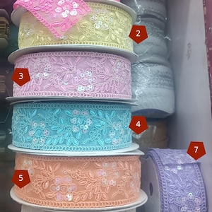 Könnte beinhalten: Ein Stapel dekorativer Bandrollen in verschiedenen Farben, darunter Rosa, Gelb, Hellblau, Pfirsich und Lavendel. Jede Rolle ist mit floralen Stickereien und Pailletten verziert, ideal für Bastelarbeiten und Verzierungen.