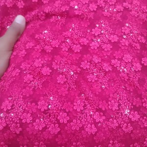 May include: Hot pink fabric with a floral pattern. The fabric is covered in small, embroidered flowers and scattered sequins. The texture appears soft and delicate, suitable for apparel or decorative projects.