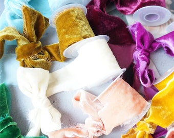 Könnte beinhalten: Sortiment von Samtbandspulen in verschiedenen Farben, darunter Türkis, Weiß, Gold, Pfirsich, Burgunderrot und Grün. Das Band wird auf Spulen mit durchsichtigen Plastikhüllen präsentiert.