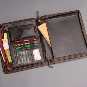 May include: A brown leather zippered portfolio with a notepad, pens, and card slots. The portfolio is open and the interior is visible.