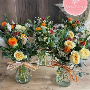 May include: Four small glass jars filled with a variety of flowers, including white roses, orange and yellow blooms, and green foliage. The jars are tied with a brown and orange ribbon. The jars are sitting on a wooden surface. The text "Barns Florals" is visible in the upper right corner of the image.