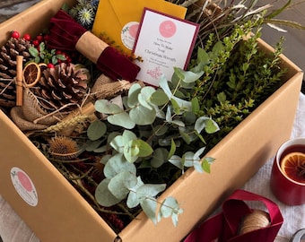 May include: A cardboard box filled with craft supplies for a Christmas wreath, including pinecones, eucalyptus branches, dried greenery, a yellow envelope, and a card titled How To Make Your Christmas Wreath. Includes red velvet ribbon and shears.