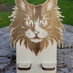 Puede incluir: Un soporte de teléfono de madera con un diseño de cara de gato grabado con láser. El soporte tiene dos ranuras para sujetar un teléfono.