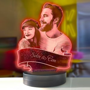 Puede incluir: Una luz nocturna LED personalizada con un retrato de pareja, delineado en neón rojo. La pancarta debajo de la imagen dice "Selin & Cem" en escritura cursiva. La luz se asienta sobre una base circular negra.