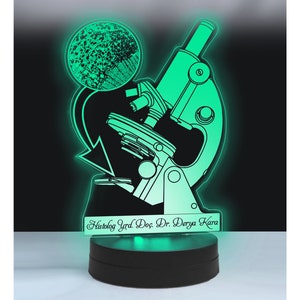 Puede incluir: Una luz nocturna 3D impresa en verde iluminada con forma de microscopio con un círculo que representa una diapositiva. La base de la luz nocturna tiene el texto "Histolog Yrd. Doc. Dr. Derya Kara".