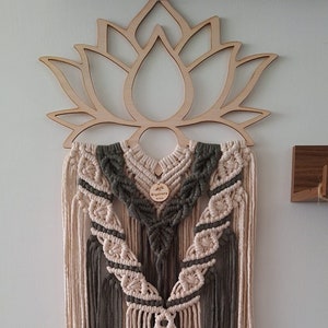May include: Macrame wall hanging featuring a lotus flower design. The wall decor has a wooden lotus flower top with a cream, olive green, and gray macrame design. The macrame has a series of knots and fringe.
