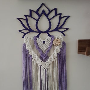 May include: A purple lotus flower wall hanging with macrame detailing. The wall decor features a wooden lotus flower outline in a deep purple hue, with white and lavender macrame threads. A small wooden tag reads "Kryations by Kyra."