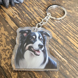 Peut inclure: Un porte-clés en acrylique transparent avec un portrait de chien Border Collie. Le chien a des poils noirs et blancs, avec une expression joyeuse. Le porte-clés a un anneau et une chaîne en métal argenté. Le fond est une surface en bois.