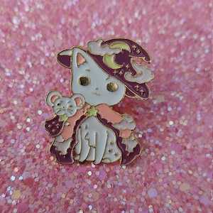 Op de afbeelding: Emaille pin met een witte kat met een paarse heksenhoed met een maan en sterren. De kat houdt een kleine muis vast, beiden in roze en paarse kleding. De pin heeft gouden accenten en staat op een roze glitter achtergrond.