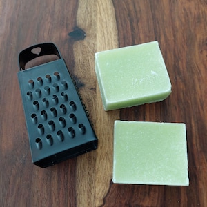May include: A black metal grater and two square, pale green soap bars are displayed on a wooden surface. The grater is on the left, and the soap is on the right. The soap has a slightly textured appearance. The wooden surface has a brown hue.