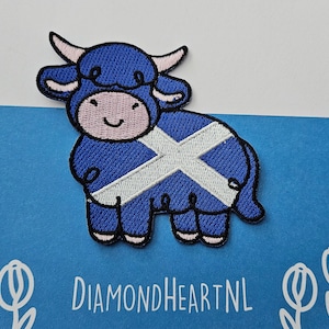 Op de afbeelding: Een blauwe geborduurde patch met een cartoon Highland-koe met een wit en blauw Schots vlagontwerp. De koe heeft roze hoorns en een lachend gezicht. De patch staat op een blauwe achtergrond met de tekst "DIAMONDHEARTNL".