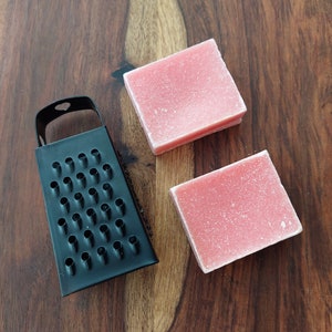 May include: A black metal grater and two square, pink soap bars sit on a dark wooden surface. The grater is rectangular with many small holes. The soap bars have a slightly textured appearance, suggesting a handmade product.