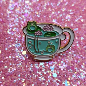 Op de afbeelding: Emaille pin met een theekopje ontwerp met een turquoise interieur en gouden rand. De theekop bevat twee kikkers, waterlelies en bellen. De theekop heeft een wit handvat en rand. De achtergrond is een roze glitter oppervlak.