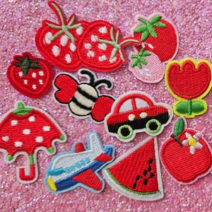 Op de afbeelding: Een verzameling geborduurde patches met diverse ontwerpen. Inbegrepen zijn aardbeien, een bij, een auto, een watermeloen, een appel, een bloem, een paraplu en een vliegtuig. De patches zijn in rood, wit, zwart, groen en blauw.