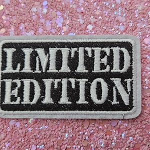 Op de afbeelding: Een rechthoekige patch met de woorden "LIMITED EDITION" in wit geborduurd op een zwarte achtergrond. De patch heeft een grijze rand en is geplaatst op een roze glitteroppervlak.