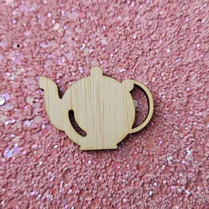 Puede incluir: Un recorte de madera con forma de tetera con asa, boquilla y tapa. La madera de color claro contrasta con el fondo de purpurina rosa. Este suministro artesanal es ideal para proyectos de bricolaje y decoraciones.