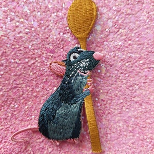 Op de afbeelding: Geborduurde patch met een cartoonrat die een gouden lepel vasthoudt. De rat heeft blauwe vacht, roze oren en neus en lacht. De achtergrond is een sprankelende roze glitter.