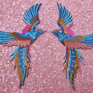 Op de afbeelding: Twee geborduurde vogelpatches met uitgespreide vleugels. De vogels hebben blauwe, bruine, roze en gouden draden. De patches liggen op een roze glitterachtergrond. Deze patches kunnen worden gebruikt voor kleding of knutselprojecten.