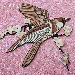 Op de afbeelding: Geborduurde patch met een vogel met uitgespreide vleugels, zittend op een bloeiende tak. De vogel heeft bruine en witte veren, met een zwarte omtrek. De bloemen zijn lichtroze, de achtergrond is glinsterend roze.