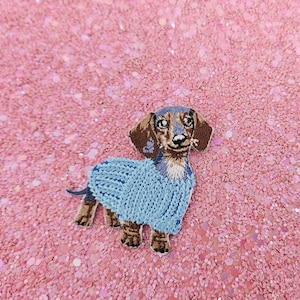 Könnte beinhalten: Gestickter Aufnäher eines Dackels mit hellblauem Pullover. Der Hund hat braunes und hellbraunes Fell mit schwarzer Nase und Augen. Der Aufnäher ist vor einem rosa, glitzernden Hintergrund angebracht.