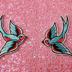 Könnte beinhalten: Zwei aufgestickte Aufnäher mit einem Schwalben-Design. Die Vögel haben türkisfarbene Flügel, rote Akzente und schwarze Umrisse. Die Aufnäher befinden sich auf einem rosa Glitzerhintergrund.