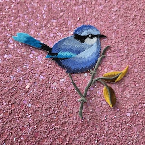 Op de afbeelding: Geborduurde patch van een blauwe vogel met zwarte accenten, zittend op een tak met gele bladeren. De vogel heeft een gedetailleerd ontwerp met verschillende tinten blauw. De achtergrond is een glinsterend roze oppervlak.