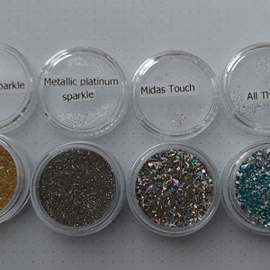 Op de afbeelding: Acht kleine, ronde, transparante potjes gevuld met verschillende glitterkleuren. De labels luiden "Pearl gold sparkle", "Metallic platinum sparkle" en "Midas Touch". De glitterkleuren omvatten goud, zilver en iriserende tinten.