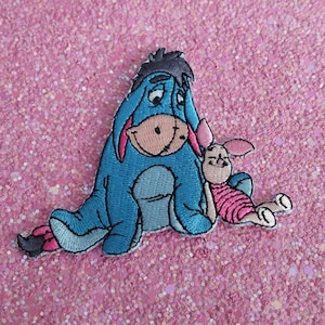 May include: Embroidered patch depicting Eeyore and Piglet from Winnie the Pooh. Eeyore is light blue with a pink inner ear, and Piglet is pink and wearing a striped shirt. The patch is on a pink glitter background.