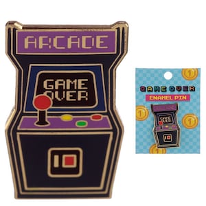 May include: Enamel pin featuring a retro arcade game design with the text "ARCADE" and "GAME OVER". The pin is black, purple, and gold with a red joystick and colorful buttons. A matching card is included.