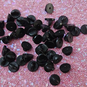 Op de afbeelding: Een verzameling zwarte pin backs verspreid op een roze glitteroppervlak. De pin backs zijn rond met een pin en een sluiting. De afbeelding toont een verscheidenheid aan pin backs, sommige met de sluiting bevestigd en sommige zonder.