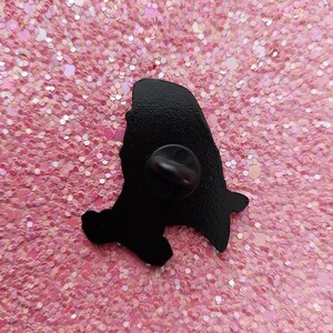 May include: A black enamel pin with a silhouette design. The pin features a rounded top and a small, pointed bottom. The pin is set against a pink glitter background.