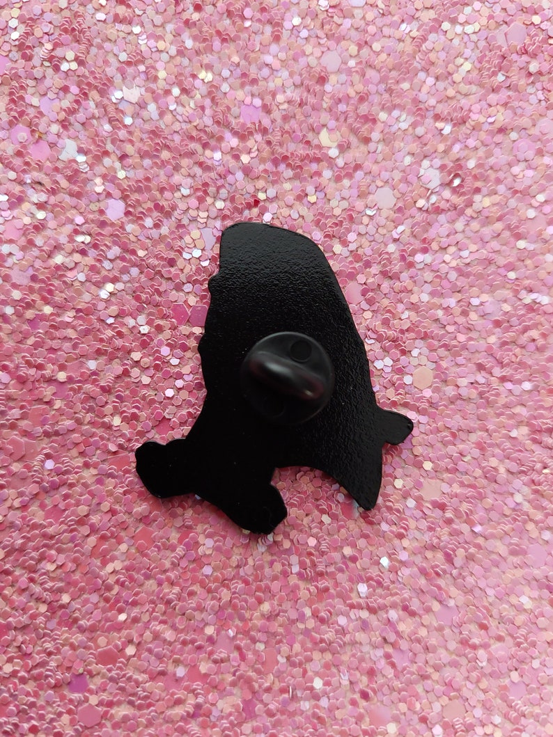 May include: A black enamel pin with a silhouette design. The pin features a rounded top and a small, pointed bottom. The pin is set against a pink glitter background.