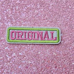 Op de afbeelding: Geborduurde patch met het woord "ORIGINAL" in roze, omlijnd in groen, op een lichtgrijze achtergrond. De patch is rechthoekig met afgeronde hoeken. De achtergrond is een roze glitteroppervlak.