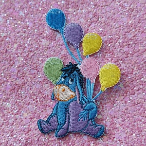 Peut inclure: Écusson brodé représentant un âne bleu tenant des ballons colorés. Le patch présente un schéma de couleurs bleu clair, violet et jaune, sur un fond rose pailleté. Idéal pour les vêtements ou les accessoires.