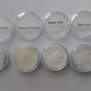 Op de afbeelding: Acht kleine, doorzichtige plastic potjes gevuld met diverse witte glitter en sparkles. De potjes zijn gelabeld met "Powdered Snow", "White Puff Twinkle", "White Glitz" en "Disco Ball". De andere potjes zijn gevuld met vergelijkbare witte glitter.