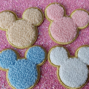 Op de afbeelding: Vier geborduurde patches in de vorm van een muizenkop. De patches zijn crème, roze, blauw en wit, elk met een gouden rand. De patches hebben een getextureerd, pluizig uiterlijk.