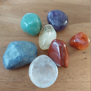 May include: Seven polished gemstones in various colors, including green, purple, orange, and clear, are arranged on a wooden surface. The stones vary in size and shape, with smooth, rounded edges. The gemstones are likely used for spiritual or decorative purposes.