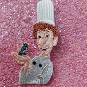 Può includere: Toppa ricamata raffigurante uno chef dei cartoni animati con cappello bianco e uniforme grigio chiaro, che tiene in mano un piccolo ratto nero e blu. Lo chef ha i capelli rossi e sorride. La toppa è su uno sfondo rosa glitterato.