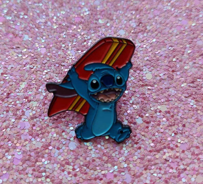 May include: A blue enamel pin featuring the character Stitch from Lilo & Stitch holding a red and yellow surfboard. The pin has a glossy finish and is set against a pink glitter background.