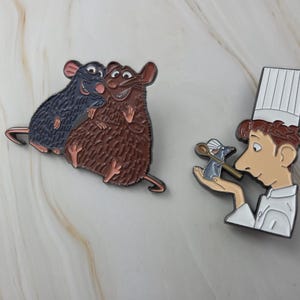 Op de afbeelding: Emaille pins met personages uit de animatiefilm. Eén pin toont twee knuffelende ratten, één blauwe en één bruine. De andere pin toont een chef-kok die een rat met een lepel vasthoudt.