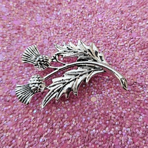 May include: A silver thistle brooch featuring intricate leaf and flower head detailing. The brooch is displayed against a pink glitter background. This decorative item is designed to be worn as an accessory, offering a touch of nature-inspired elegance. The brooch is a classic design.