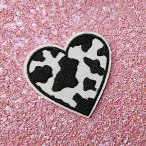 Op de afbeelding: Een hartvormige patch met een koeienprint ontwerp. De patch heeft een witte achtergrond met zwarte vlekken, omlijnd met een zwarte rand. De patch is geplaatst tegen een roze glitterachtergrond.