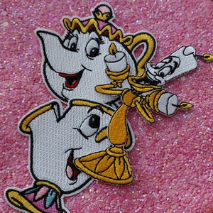 Könnte beinhalten: Gestickte Aufnäher mit Figuren aus dem Zeichentrickfilm. Die Aufnäher zeigen eine weiße Teekanne mit goldener Krone und lächelndem Gesicht sowie einen goldenen Kerzenhalter mit Kerze. Die Aufnäher befinden sich auf einem rosa Glitzerhintergrund.