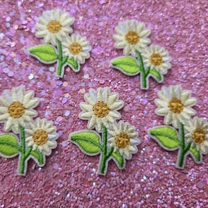 Puede incluir: Parches bordados de margaritas con pétalos blancos y centros amarillos, combinados con hojas y tallos verdes. Los parches están dispuestos sobre una superficie rosa brillante, perfectos para manualidades o para adornar ropa.