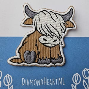 Op de afbeelding: Geborduurde patch met een cartoon Highland-koe in bruin, beige, grijs en wit. De koe zit met haar hoofd naar beneden. De patch heeft een witte rand en de tekst "DIAMONDHEARTNL" is zichtbaar.