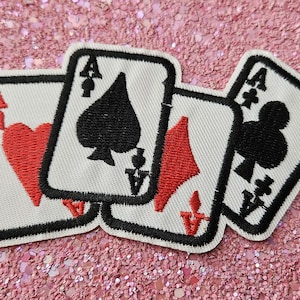 May include: Four playing card patches featuring the Ace of Hearts, Spades, Diamonds, and Clubs. Each card has a white background with black borders and suit symbols. The patches are on a pink glitter surface.
