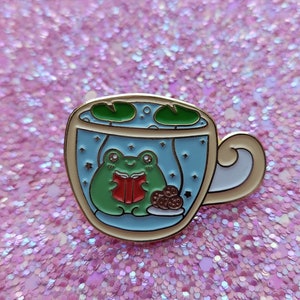 Op de afbeelding: Emaille pin met een groene kikker die een rood boek leest in een theekopje. De theekop heeft een blauwe binnenkant met waterlelies en sterren. Een bord met koekjes staat onderaan. De pin heeft een gouden omtrek en een wit handvat.