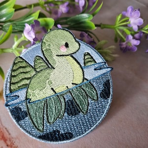 Pode incluir: Remendo bordado com um dinossauro de desenho animado nadando na água. O dinossauro é verde claro com um rubor rosa, e a água é azul. O remendo é circular com uma borda azul e um fundo texturizado.