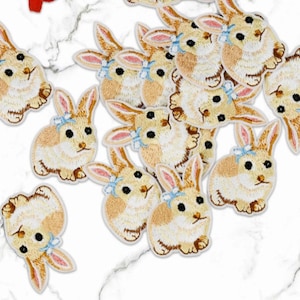 Op de afbeelding: Geborduurde konijnenpatches in crème, beige en roze met blauwe strikken. Deze patches hebben gedetailleerde stiksels en zijn verspreid over een wit marmeren oppervlak. Ideaal om een decoratieve touch toe te voegen aan kleding of accessoires.