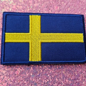 Puede incluir: Un parche rectangular con la bandera de Suecia. El parche tiene un fondo azul oscuro con una cruz nórdica amarilla. La cruz se extiende hasta los bordes del parche. El parche es probablemente de tela y tiene un borde cosido.